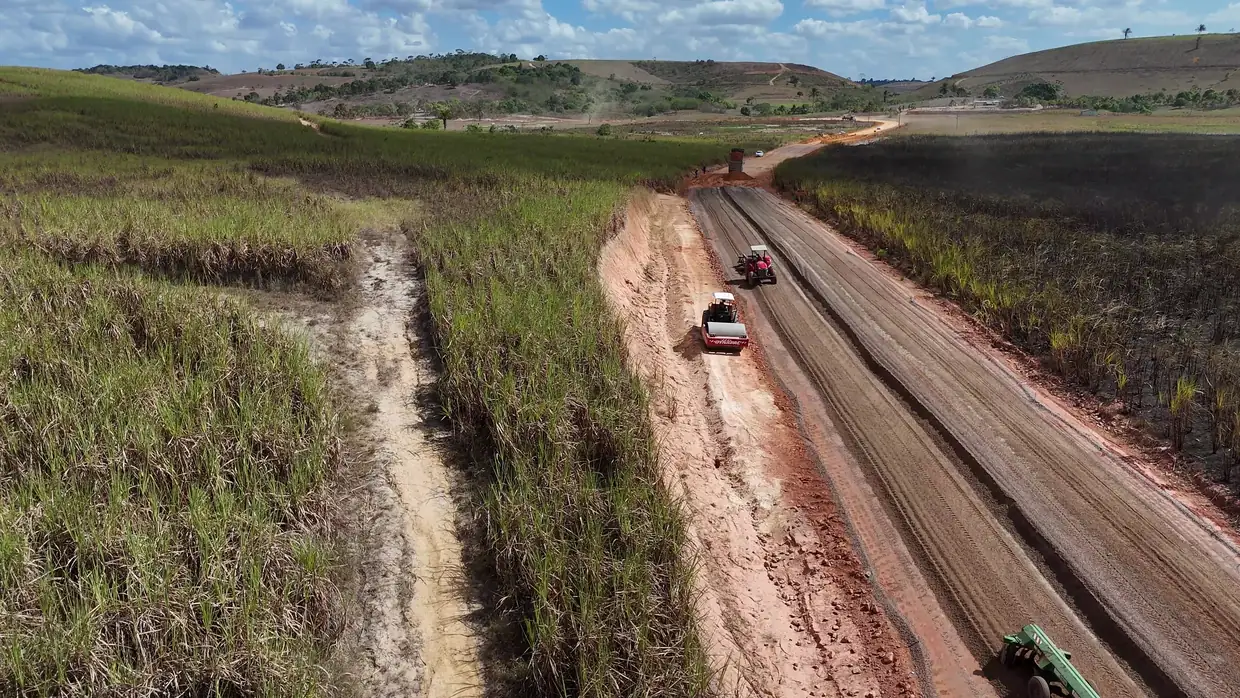 Caminhões na estrada do Acesso Onça em obras, entre canaviais, vistos do alto